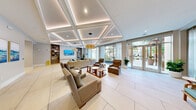 Located at 1305 Morgan Stanley Ave in Winter Park, Florida, this single-floor commercial lobby and lounge space offers a generously scaled, thoughtfully designed interior that makes a strong first impression. The space is defined by a dramatic double-height atrium entrance, where floor-to-ceiling glass walls flood the entry with natural light and provide open views of the surrounding palm-lined streetscape.

A signature curved teal feature wall anchors the central foyer, paired with a built-in display shelf and seating area that adds both visual impact and functional warmth. Sculptural pendant light fixtures in warm gold tones hang overhead, complementing the bright, airy palette throughout.

The adjacent lounge area features an elegant coffered ceiling with integrated ambient lighting, creating a refined and comfortable gathering space. Large-format porcelain tile flooring runs continuously throughout, enhancing the sense of openness. Generous seating arrangements with neutral upholstered furnishings and teal accents invite relaxed interaction, while sheer floor-to-ceiling drapery softens the abundant natural light from expansive glass doors leading outdoors.

A dedicated corridor leads to additional private rooms and service areas, accented with curated artwork. The Juno-branded reception area is visible from the main hall, reinforcing the professional, polished character of this well-appointed lobby environment.