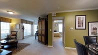 This single-floor residence presents a welcoming interior defined by warm, earthy color tones and comfortable, carpeted flooring throughout. The main living area is accented by olive-green walls that add depth and character to the space, complemented by crisp white crown molding that frames the ceiling. A ceiling fan provides both function and a classic aesthetic touch, while large windows dressed with traditional valances invite abundant natural light, brightening the room considerably.

A substantial dark wood armoire serves as a prominent furniture piece, adding a traditional charm to the living area. Decorative greenery atop the armoire and a classical column-style plant stand near the windows enhance the warm, homey atmosphere. A patterned area rug anchors the space, introducing additional texture underfoot.

An open doorway connects the living area directly to the bedroom, which features similarly carpeted flooring, neutral tones, and its own window allowing natural light to filter through. The bedroom is furnished with a bed dressed in warm-toned bedding, contributing to the cohesive, comfortable feel of the overall residence. The layout flows naturally between rooms, creating an open and accessible single-level living experience.