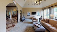 This single-floor space showcases a refined Mediterranean-inspired aesthetic, characterized by graceful arched doorways, soaring vaulted ceilings, and warm golden-toned walls finished with a textured plaster treatment. The main lounge area is generously proportioned, anchored by a patterned area rug in soft blue tones that complements the neutral carpeting underfoot. Comfortable seating arrangements, including upholstered sofas and armchairs with decorative accent pillows, create an inviting atmosphere for gathering and relaxation. A wall-mounted flat-screen television is flanked by framed artwork, while a statement chandelier with a wrought iron ring design adds a dramatic focal point overhead. Decorative ironwork gates and tall sculptural vases lend an artistic, Old World charm to the space. Large windows dressed with linen drapery flood the interior with natural light and offer views of lush greenery outside. Arched openings with classical columns connect the main lounge to adjacent areas, including a tiled transitional entry and a secondary room visible beyond, hinting at additional functional spaces. Potted topiaries and carefully curated décor reinforce the cohesive, upscale design throughout.