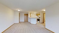 This single-floor unit offers an open-concept living and kitchen layout that maximizes the sense of space throughout. The living area is finished with plush, light-toned carpeting and neutral white walls, creating a clean and welcoming atmosphere. Flush-mounted ceiling lights provide consistent illumination across the space.

The kitchen is positioned centrally within the open floor plan, anchored by a peninsula counter that serves as both a functional workspace and a natural divider between the kitchen and living area. Warm honey-toned wood cabinetry lines the kitchen walls, complementing the white appliances, which include a range, microwave, and refrigerator. A pendant light hangs above the peninsula, adding a subtle design accent.

A wood-paneled entry door marks the main entrance, with an additional interior door nearby providing access to a bathroom or utility space. The overall layout is efficient and practical, with clear sightlines throughout.

Externally, the building is part of a well-maintained multi-story complex set within a landscaped environment featuring rolling green lawns, mature evergreen trees, a decorative pond, and open green spaces, contributing to a tranquil, park-like setting.