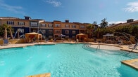 Located at 115 Amberleigh Dr in Wilmington, NC, this single-floor residence is part of a well-appointed multi-building community designed with a resort-inspired aesthetic. The exterior architecture features a combination of warm tan, navy blue, and amber-toned siding accented by shuttered windows, creating a visually cohesive and attractive streetscape framed by mature palm trees and lush landscaping.

The unit includes a covered private patio finished with stone-tile flooring, providing a comfortable outdoor retreat with views of manicured greenery, ornamental iron fencing, and the surrounding community grounds. The patio is furnished with wicker seating and a bistro-style table, making it well-suited for relaxing or casual outdoor dining.

Community amenities are a prominent feature of this property. A generously sized resort-style swimming pool serves as the centerpiece of the outdoor common area, surrounded by a spacious pool deck with lounge chairs, shaded cabanas, and umbrella-covered seating. An outdoor entertainment station features a granite countertop bar with a built-in sink, mounted flat-screen television, and wicker bar seating—ideal for social gatherings. Additional seating areas with bistro tables are positioned throughout the courtyard, all overlooking the pool and lush palm-lined surroundings under the sunny Wilmington sky.