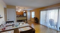 This single-floor space features an open-concept design that seamlessly integrates the kitchen, dining, and living areas into one cohesive layout. The kitchen is equipped with wooden cabinetry in a warm honey tone, a dark countertop breakfast bar with bar stool seating, and standard appliances including a refrigerator. Pendant lights hang above the kitchen area, providing focused task lighting that complements the overall warm ambiance of the space.

The dining area accommodates a dark wood dining table with seating for multiple guests, dressed with decorative placemats that add a homey touch. Parquet-style flooring in a light natural wood tone extends throughout the open area, adding texture and visual interest underfoot.

Natural light is a standout feature of this space, with a large sliding glass door fitted with vertical blinds allowing generous light to pour in, along with an additional window that further brightens the interior. A wine rack near the sliding door adds a functional decorative element. The warm yellow-toned walls contribute to the inviting and comfortable atmosphere of the space.