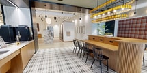 Located at 415 N Lake St in Madison, Wisconsin, this 510-square-foot single-floor lobby and lounge space delivers a bold, contemporary design aesthetic with a wealth of distinctive architectural details. The floor is covered in a striking black-and-white plaid tile pattern that anchors the room's eclectic character, transitioning seamlessly to polished concrete near the reception area. A curved, fluted wood bar with black spindle chairs serves as a central gathering point, illuminated overhead by a dramatic double-tiered chandelier featuring amber glass accents. The reception area showcases a substantial marble-clad desk with built-in mailboxes, set against a vibrant multicolored checkered accent wall and complemented by globe pendant lights. A plush red tartan upholstered banquette along one wall provides additional seating, paired with round marble-top café tables. Large windows allow natural light to filter in, offering views of the surrounding greenery. Tall tropical plants add organic warmth to the space. The service counter area features warm wood cabinetry, modern point-of-sale equipment, and sleek pendant lighting. A dark metal elevator surround adds an industrial contrast. Together, these elements create a vibrant, welcoming communal environment with a refined yet playful design sensibility.