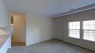 This single-story space centers around a spacious living area designed with a clean, neutral aesthetic. The room is finished with wall-to-wall carpet in a light beige tone, contributing to a warm and understated atmosphere throughout. Crisp white walls and a smooth ceiling with a subtle beam detail provide a simple architectural framework, while a ceiling-mounted sprinkler and a visible HVAC vent indicate modern building utilities integrated into the design.

A large window adjacent to the front entry door allows natural light to fill the space, fitted with blinds for adjustable privacy and light control. The white paneled entry door serves as the main access point, positioned at the far end of the living area. A thermostat is mounted on the wall, offering convenient climate control.

A hallway extends from the living area, leading to additional areas of the unit. A louvered closet door is visible along the hallway wall, providing dedicated storage space. The overall layout is open and straightforward, making it a functional and adaptable living environment with a neutral palette suited to a variety of furnishing styles.