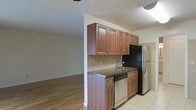 This single-floor space offers a functional and open layout, highlighted by a well-appointed kitchen that flows seamlessly into an adjacent living area. The kitchen features raised-panel cabinetry in a warm medium-brown finish, providing ample upper storage. Dark granite countertops add a rich contrast and a sense of durability, while a tile backsplash in neutral tones frames the workspace neatly. Stainless steel appliances, including a refrigerator and dishwasher, complement the kitchen's practical design. The sink is positioned along the main counter run, offering a clean and efficient workspace. The kitchen floor transitions from tile to the warm hardwood flooring that defines the open living area beyond. A structural column subtly separates the two spaces while maintaining the open feel. The living area benefits from generous natural-feeling light and smooth walls, creating a clean, unfussy backdrop. Overall, the space combines functional kitchen design with an open, connected living area, making it a straightforward and practical single-floor layout.