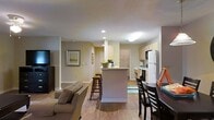 This thoughtfully designed open-concept space seamlessly integrates kitchen, dining, and living areas, creating a harmonious and functional environment. The heart of the space is a kitchen with light-colored cabinetry and modern appliances, including a white refrigerator. A peninsula with bar seating serves as a natural divider between the kitchen and living area, enhancing the flow of the space.

The living room features comfortable seating, with a neutral-toned sofa visible in the foreground. Hardwood flooring extends throughout, adding warmth and continuity. Recessed lighting in the ceiling provides even illumination, while artwork on the walls adds visual interest.

A dining area is situated adjacent to the kitchen, furnished with a dark wood table and chairs, perfect for casual meals or entertaining. The neutral wall color creates a calm backdrop, allowing for versatile decorating options. Overall, this space embodies a contemporary aesthetic with its open layout and cohesive design elements.