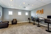 Situated at 1401 College Ave in Manhattan, Kansas, this single-floor commercial space offers a well-appointed and functional environment designed to meet a variety of professional needs. The interior is finished with a consistent palette of cool gray walls, neutral-toned carpet tiles, and suspended drop ceilings with recessed lighting throughout, creating a clean and cohesive aesthetic.

The space includes a dedicated business center equipped with multiple computer workstations set along wall-mounted desks, accompanied by adjustable bar-height seating. Decorative wood panel wall art adds a warm, modern accent to this area. Compact square windows positioned along the walls allow natural light to filter in while maintaining a focused work atmosphere.

A comfortable lounge area features dark leather armchairs arranged for relaxed seating, accented by a large decorative wall clock that adds character to the space. This area also benefits from the same square window design, providing a consistent architectural detail across the floor.

An open corridor connects the various zones, revealing a reception or service counter area visible in the background, suggesting the space functions as a shared-use or community-oriented facility. The layout flows intuitively between workspaces and seating areas, making the overall environment both practical and welcoming for everyday use.
