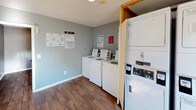 This single-story laundry facility is designed to serve shared-use needs efficiently. The space is organized across two connected rooms, each finished with durable wood-style plank flooring and neutral-toned walls that create a clean, functional environment. The textured ceilings complement the straightforward, practical aesthetic throughout.

The main laundry room houses a row of three top-load washing machines, each equipped with digital payment terminals accepting card transactions, making cashless use convenient. Instructional and service signage is posted prominently on the walls, providing users with clear guidance on machine operation and maintenance contacts. Stacked washer and dryer units are positioned along one wall, maximizing the use of vertical space and expanding overall laundry capacity.

An adjoining anteroom serves as an entry buffer, featuring a large window that allows natural light to fill the space. Its light-colored walls and open layout provide a comfortable transition into the laundry area. A doorway connects the two rooms, facilitating smooth movement between them.

Overall, the facility is practical and well-organized, with adequate natural light, modern payment-enabled machines, and a tidy layout designed to accommodate regular, high-volume use.