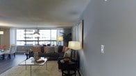 This single-floor space offers a thoughtfully arranged living area that combines comfort and contemporary style. The living room is the centerpiece of the space, characterized by expansive windows that flood the room with natural light and offer open views of the surrounding urban landscape. The neutral gray walls provide a calm, versatile backdrop that complements the modern décor throughout.

The living area is generously proportioned, comfortably accommodating a large, cushioned sectional sofa dressed with decorative throw pillows in warm tones. A glass-topped coffee table anchors the seating area, while a small accent table and chair create a cozy secondary nook. Layered lighting adds depth and warmth to the room, with a sleek arc floor lamp and multiple table lamps casting a soft, inviting glow.

A large piece of framed artwork on the accent wall adds a bold, artistic focal point, while a lush indoor plant near the windows introduces a touch of natural greenery. The room is finished with a plush area rug that defines the seating zone and adds texture underfoot. Overall, the space presents a balanced blend of functionality and contemporary design, making it a comfortable and visually appealing environment.