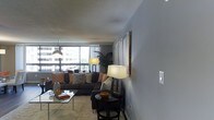 This single-floor space offers a thoughtfully arranged living area that combines comfort and contemporary style. The living room is the centerpiece of the space, characterized by expansive windows that flood the room with natural light and offer open views of the surrounding urban landscape. The neutral gray walls provide a calm, versatile backdrop that complements the modern décor throughout.

The living area is generously proportioned, comfortably accommodating a large, cushioned sectional sofa dressed with decorative throw pillows in warm tones. A glass-topped coffee table anchors the seating area, while a small accent table and chair create a cozy secondary nook. Layered lighting adds depth and warmth to the room, with a sleek arc floor lamp and multiple table lamps casting a soft, inviting glow.

A large piece of framed artwork on the accent wall adds a bold, artistic focal point, while a lush indoor plant near the windows introduces a touch of natural greenery. The room is finished with a plush area rug that defines the seating zone and adds texture underfoot. Overall, the space presents a balanced blend of functionality and contemporary design, making it a comfortable and visually appealing environment.