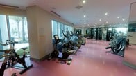 This fitness center offers a comprehensive workout environment thoughtfully designed to accommodate a variety of exercise routines. The space is lined with large floor-to-ceiling windows along one wall, flooding the interior with natural light and providing an open, energizing atmosphere. A row of modern cardio machines — including stationary bikes, ellipticals, and treadmills — is neatly arranged along the windowed side, offering an inspiring backdrop for endurance training. The distinctive rubberized flooring in a warm pink-red tone provides both safety and comfort underfoot throughout the space. Toward the rear, a dedicated strength training zone houses a robust selection of resistance machines and free weights, complemented by mirrored walls that enhance the sense of depth and allow users to monitor their form. Recessed ceiling lighting works in harmony with the natural light to ensure the entire area is well-illuminated at all times. The layout is spacious and organized, allowing for easy movement between equipment zones. Clean lines, a neutral color palette on the walls, and modern equipment selections give the facility a polished, professional appearance suited for both casual and dedicated fitness enthusiasts.