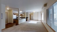 This well-designed single-floor residence offers an open-concept layout that seamlessly connects the kitchen and living areas, creating a sense of expansive, flowing space. The kitchen is outfitted with warm wood cabinetry, dark countertops, stainless steel appliances including a full-size refrigerator and dishwasher, and a built-in cooktop. Pendant lighting hangs above the kitchen counter, adding both functional illumination and a modern decorative touch. A structural column subtly defines the boundary between the kitchen and the adjoining living space without interrupting the open feel. The living area is finished with plush, light-toned carpeting that adds warmth underfoot, while natural light streams in through a large window, brightening the room throughout the day. Flush-mount ceiling fixtures provide consistent ambient lighting. A hallway visible toward the rear of the space leads to additional rooms, suggesting a thoughtful floor plan that balances communal and private areas. Clean white walls and neutral tones throughout contribute to a calm, inviting atmosphere that is both versatile and easy to personalize.