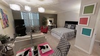 This thoughtfully designed studio with one bathroom offers a seamless blend of functionality and contemporary style. The open-concept layout maximizes the available space, integrating the sleeping and living areas into one cohesive environment. The main area is anchored by a full-sized bed dressed in a chevron-patterned comforter, complemented by a dark upholstered headboard. An accent wall in a warm charcoal tone serves as a striking focal point, adding depth and contrast to the otherwise light and airy color palette. Plush carpeting runs throughout, contributing to a comfortable and welcoming ambiance. The living area features a sofa and a patterned accent chair, creating a cozy seating arrangement. A sleek glass coffee table with vibrant pink accents adds a bold pop of color, enhancing the modern aesthetic. A tall arc floor lamp and a crystal-style pendant light fixture provide both task and ambient lighting, elevating the space's overall design. Teal and white chevron curtains frame the windows, allowing natural light to filter in while tying together the room's cohesive décor theme. A vanity desk and a lively red accent chair complete the space, offering both utility and visual interest throughout.