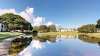 This single-story residence is situated within a beautifully maintained Florida community, offering a tranquil setting defined by its natural surroundings and thoughtful landscaping. The exterior presents a classic Florida architectural style, characterized by a neutral palette, a pitched roof, and a low-profile design that blends seamlessly into the lush environment.

The property is nestled alongside a calm, reflective pond that mirrors the sky and surrounding tropical canopy, creating a picturesque waterfront backdrop. Expansive stretches of manicured green lawn extend from the building's perimeter to the water's edge, providing a sense of openness and connection to nature. Mature oak trees, palms, and dense tropical vegetation frame the grounds, offering shade and a layered, verdant landscape throughout.

The community setting enhances the appeal of the space, with well-kept pathways and carefully tended garden beds contributing to an overall atmosphere of serene, resort-style living. The abundance of natural light and open skies typical of the Florida climate further elevate the outdoor experience. Whether enjoyed from the exterior or viewed from within, the sweeping water and garden vistas offer a consistently peaceful and scenic environment that defines the character of this property.