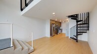This contemporary two-story loft-style space is defined by clean lines, an open layout, and modern finishes throughout. The main level features light hardwood flooring that extends across the open living area and into the kitchen, creating a seamless, cohesive aesthetic. The kitchen is well-appointed with warm wood-toned cabinetry, a gray tile backsplash, dark countertops, and a full suite of stainless steel appliances including a refrigerator, range, microwave, and dishwasher. A compact, modern bathroom on the main level includes a floating wood-trimmed vanity with a dark countertop, an undermount sink, and a backlit mirror, alongside a separate tub-shower combo with built-in shelving. A striking staircase with a mix of black metal railings and light wood accents connects the two levels. The upper floor offers an open loft area with the same light hardwood flooring, enhanced by large windows that bring in abundant natural light and provide views of the surrounding area. The space's minimalist design, neutral color palette of white walls and natural wood tones, and thoughtful architectural details such as the open mezzanine-style upper level contribute to a bright, airy, and functional living environment.