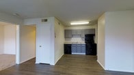 This single-floor space presents an open-concept layout that seamlessly connects the living area to the kitchen. The kitchen is outfitted with gray lower and upper cabinetry, a light-toned countertop, and a full suite of black appliances, including a range, overhead exhaust hood, and dishwasher. A stainless sink is positioned beneath upper cabinets, providing a functional and organized workspace. Overhead fluorescent lighting ensures the kitchen is well-lit for everyday use.

The flooring throughout the space features wood-style planks in a warm, medium-toned finish, adding a cohesive and contemporary feel. The walls are finished in a neutral off-white, contributing to a clean and airy atmosphere. Ceiling-mounted HVAC vents indicate a central air system, while white baseboards frame the perimeter of each room neatly.

A white interior door with a brass knob leads to an adjacent room or hallway, suggesting additional living spaces beyond the main area. The layout feels practical and efficient, with clear sightlines from the living space to the kitchen, making it well-suited for everyday functionality.