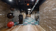 Located at 231 Bridgeway Circle in Nashville, TN, this fitness center offers a comprehensive single-floor exercise facility designed to support a full range of workout routines. The space features a striking industrial-modern aesthetic, highlighted by an exposed black ceiling with visible ductwork and ductwork, bold circular overhead lighting, and a combination of natural brick accent walls and smooth concrete-finish surfaces that lend the space a contemporary, urban character.

The exercise area is thoughtfully organized into distinct workout zones. A dedicated cardio section lines one side of the room, equipped with treadmills, elliptical trainers, and stationary cycles by True Fitness, complemented by wall-mounted televisions for entertainment during workouts. Opposite, a strength training zone features a selection of selectorized cable and weight machines alongside a free weight rack stocked with a full range of dumbbells, all reflected in full-length mirrors that amplify the sense of space. A separate stretching and functional training area is finished with hardwood flooring and houses medicine balls and other accessories.

The flooring throughout the main exercise space consists of a teal, gray, and white modular carpet tile pattern that adds a vibrant visual contrast to the darker ceiling and walls. Glass partitions define the space's entry zone, creating a clean, open transition between areas. The overall ambiance is energetic and professional, making this fitness center a well-rounded facility for residents and users alike.