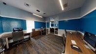 This single-floor office suite is designed to support a functional and collaborative work environment, organized into two primary interconnected zones. The space is defined by a striking two-tone wall treatment, with bold cobalt blue on the lower half and soft gray above, separated by clean white chair rail molding — a consistent design element that gives the entire suite a cohesive, professional appearance.

The main workspace accommodates multiple workstations featuring warm wood-toned desks paired with ergonomic task chairs. Low glass privacy panels divide individual work areas while maintaining an open, communicative atmosphere. Workstations are equipped with monitors and supporting peripherals, making the space immediately functional. Linear LED light fixtures are suspended from the ceiling, providing consistent, well-distributed illumination throughout.

A glass-walled partition with a sliding door separates the main workspace from a dedicated conference or meeting room. This enclosed area features a light wood conference table surrounded by upholstered chairs, offering a private setting for meetings or focused collaboration.

Patterned carpet tiles in dark tones with blue striping run throughout, complementing the color scheme and adding texture underfoot. Windows fitted with blinds allow natural light to filter into the space, contributing to a balanced and practical office environment.