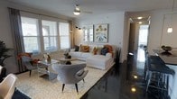 This single-floor space presents a thoughtfully designed living area that balances modern aesthetics with comfortable functionality. The living room is anchored by striking polished dark concrete floors that contrast elegantly with the bright white walls, creating a sophisticated visual dynamic throughout the space.

The living area is generously proportioned, accommodating a large white sectional sofa dressed with an array of decorative pillows in warm amber, navy, and neutral tones. A coordinating grey accent chair with mid-century modern styling sits opposite, centered on a plush cream shag area rug that adds warmth and texture underfoot. A sleek acrylic coffee table completes the seating arrangement, contributing to the contemporary feel.

Natural light flows into the space through multiple windows fitted with white horizontal blinds and floor-length curtains, keeping the atmosphere bright and airy. A ceiling fan with a chrome finish provides both practical ventilation and a clean design element overhead. Two framed botanical art prints add a refined decorative touch to the walls. A tall arched floor lamp and potted snake plant introduce additional design layers. A hallway visible from the living area suggests further rooms beyond.