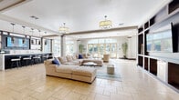 Located at 940 W Canton Ave in Winter Park, Florida, this sophisticated single-floor clubhouse lounge offers a generously sized, open-plan gathering space designed for both comfort and style. The entrance welcomes visitors with soaring arched windows, decorative columns, and an abundance of natural light that pours in from floor-to-ceiling glass doors overlooking lush outdoor surroundings. Tall, tropical potted plants and elegant accent chairs create a refined yet inviting first impression.

The expansive living room anchors the space with a large sectional sofa dressed in neutral tones and navy accents, arranged atop a contemporary striped area rug. Warm travertine stone tile flooring flows seamlessly throughout, complementing the bright, airy atmosphere. Overhead, a pair of bold drum pendant chandeliers provide ambient illumination alongside recessed lighting.

A sleek wet bar and kitchen station lines one wall, featuring dark cabinetry, stainless steel appliances, a long white countertop with bar seating, and cascading pendant lights — perfect for hosting and entertaining. A striking dark wood open-shelving unit with an integrated flat-screen television serves as a sophisticated focal point, dividing the lounge from additional seating areas. Expansive glass doors throughout the space connect the interior to outdoor views, reinforcing the bright, Florida-inspired ambiance.