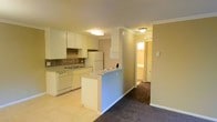 This single-floor space showcases a functional and open layout, connecting the kitchen and living areas in a cohesive design. The kitchen is well-appointed with white cabinetry, a tiled backsplash, white appliances including a refrigerator and dishwasher, and a double sink positioned beneath upper cabinets. Light tile flooring runs throughout the kitchen, providing a clean, bright foundation. A peninsula with a raised breakfast bar serves as a natural divider between the kitchen and the adjacent living area, offering additional counter space and a casual seating option.

Crown molding accents the ceilings throughout, adding a polished architectural detail to the space. The living area transitions seamlessly from the kitchen, featuring dark carpeting that adds warmth and contrast. Flush-mount ceiling fixtures provide consistent overhead lighting across the space.

A hallway visible from the main living area leads to additional rooms, with a glimpse of a naturally lit bathroom at the far end, suggesting a practical and well-organized floor plan. The neutral color palette of beige and white walls creates a versatile backdrop throughout the space.