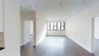 This single-floor space showcases a clean, contemporary design characterized by a neutral, all-white color palette that enhances the sense of openness and airiness throughout. The main living area is anchored by rich, dark hardwood flooring that contrasts beautifully with the crisp white walls and ceiling, creating a sophisticated yet inviting atmosphere. Recessed lighting is thoughtfully distributed across the ceiling, providing even, modern illumination throughout the space. A large, multi-pane window with dark-framed casings dominates one wall of the main area, welcoming an abundance of natural light and offering views of the surrounding urban environment. The clean white baseboards and smooth wall finishes reflect the space's attention to detail and quality of construction. An open doorway leads to an adjacent room, also finished with the same hardwood flooring and white walls, suggesting a consistent and cohesive design throughout. The layout flows naturally between spaces, offering a versatile and functional floor plan. The overall ambiance is bright, minimal, and polished, making it well-suited for a variety of uses.