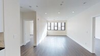 This single-floor space showcases a clean, contemporary design characterized by a neutral, all-white color palette that enhances the sense of openness and airiness throughout. The main living area is anchored by rich, dark hardwood flooring that contrasts beautifully with the crisp white walls and ceiling, creating a sophisticated yet inviting atmosphere. Recessed lighting is thoughtfully distributed across the ceiling, providing even, modern illumination throughout the space. A large, multi-pane window with dark-framed casings dominates one wall of the main area, welcoming an abundance of natural light and offering views of the surrounding urban environment. The clean white baseboards and smooth wall finishes reflect the space's attention to detail and quality of construction. An open doorway leads to an adjacent room, also finished with the same hardwood flooring and white walls, suggesting a consistent and cohesive design throughout. The layout flows naturally between spaces, offering a versatile and functional floor plan. The overall ambiance is bright, minimal, and polished, making it well-suited for a variety of uses.