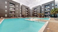 This multi-story residential complex is built around a striking U-shaped courtyard that serves as the centerpiece of its outdoor amenity experience. The architecture features a contemporary design with dark horizontal lap siding, bold red accent trim around windows and entryways, and private balconies that overlook the courtyard on every floor — spanning four stories above the ground level.

The focal point of the outdoor space is a generously sized resort-style swimming pool with vivid turquoise water, complemented by built-in in-pool lounge chairs and a tanning ledge for relaxed sunbathing. Adjacent to the main pool is a separate spa or wading pool, clearly marked with depth indicators, providing an additional relaxation option. The pool deck is finished in paver-style brick, adding a refined texture underfoot.

A pergola structure anchors one corner of the deck, shading a dedicated outdoor lounge area furnished with plush red sofas and dark wicker seating, creating an inviting social gathering space. Additional chaise lounge chairs are arranged along the pool's perimeter, offering ample space for sunbathing. Red accent chairs are also positioned at various points around the deck, tying the color scheme together cohesively.

The entire courtyard is enclosed by the surrounding building wings, offering residents a sense of privacy and security while maintaining an open-air atmosphere ideal for outdoor living.