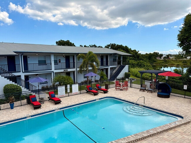 Encantador complejo de apartamentos con una pintoresca zona de piscina con tumbonas rojas, sombrillas y un exuberante paisaje verde de fondo. - Waterside Village