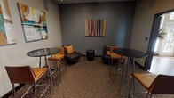 This well-appointed single-floor lounge space is thoughtfully designed to accommodate both casual socializing and informal gatherings. The space is divided into complementary zones, each offering a distinct seating experience. A cozy conversation area features dark leather swivel chairs accented with mustard-yellow throw pillows, arranged around a sleek black accent table. The warm-toned patterned carpeting grounds the space and adds texture underfoot throughout.

Bistro-height round tables paired with contemporary bar-style chairs provide additional seating options for those seeking a more upright setting. A patterned upholstered ottoman serves as an additional accent piece, enhancing the layered, curated aesthetic of the lounge.

The walls are finished in a sophisticated slate-gray tone, serving as a gallery-like backdrop for a rotating collection of bold, colorful abstract paintings that add vibrancy and visual interest throughout. A built-in beverage station with dark cabinetry and granite-style countertops, complete with a commercial-grade coffee machine, offers convenient refreshment access.

A wall-mounted flat-screen television anchors one side of the space, adding an entertainment element. An adjacent room, visible through a wide doorway, provides overflow seating and features natural light from large windows, a fireplace, and hardwood flooring, extending the overall warmth and functionality of the space.