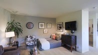 This single-floor space presents a compact yet well-appointed living area designed with both comfort and personality in mind. The room is finished with neutral gray and beige wall tones that create a calm, cohesive backdrop for the eclectic furnishings throughout. Light-colored carpeting runs underfoot, contributing to the room's warm and inviting ambiance.

The living area centers around a daybed-style sofa dressed in a gray patterned cover and accented with a mix of decorative pillows in earthy tones and a pop of orange, adding visual interest. Adjacent to it sits a distinctive black-and-white cowhide patterned accent chair, introducing a bold design element to the space.

A dark wood media console anchors one wall, supporting a large flat-screen television and decorative accessories including a globe, lending a well-traveled aesthetic. A coordinating desk lamp provides ambient lighting. The walls are adorned with an eclectic gallery arrangement of framed artwork and maps, alongside a large round decorative wall clock, adding character and depth. A small wooden tray with plants and candles on the floor adds a natural, lived-in finishing touch.