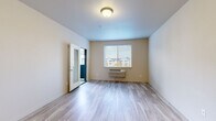 This single-floor residential unit offers a clean, contemporary design with a thoughtful layout and consistent finishes throughout. The main living area features light-toned luxury vinyl plank flooring, neutral wall colors, and a window fitted with horizontal blinds that frames views of the neighboring community, allowing natural light to fill the space. A wall-mounted HVAC unit provides climate control, and a glazed interior door adds a touch of modern detailing.

The kitchen is a standout feature, showcasing rich dark-wood cabinetry in a warm walnut finish paired with crisp white countertops. Stainless steel appliances — including a refrigerator, range, and over-the-range microwave — lend a polished, functional appeal, while pendant lighting above adds an elegant finishing touch.

The bathroom is finished with matching dark-wood cabinetry, a white countertop vanity, and wood-look vinyl flooring. A full soaking tub with shower completes the space. The bedrooms are carpeted for comfort, with large windows offering pleasant outdoor views and an abundance of natural light. An in-unit laundry area is also present, adding everyday convenience. Overall, the unit presents a cohesive, modern aesthetic well-suited for comfortable residential living.