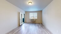 This single-floor residential unit offers a clean, contemporary design with a thoughtful layout and consistent finishes throughout. The main living area features light-toned luxury vinyl plank flooring, neutral wall colors, and a window fitted with horizontal blinds that frames views of the neighboring community, allowing natural light to fill the space. A wall-mounted HVAC unit provides climate control, and a glazed interior door adds a touch of modern detailing.

The kitchen is a standout feature, showcasing rich dark-wood cabinetry in a warm walnut finish paired with crisp white countertops. Stainless steel appliances — including a refrigerator, range, and over-the-range microwave — lend a polished, functional appeal, while pendant lighting above adds an elegant finishing touch.

The bathroom is finished with matching dark-wood cabinetry, a white countertop vanity, and wood-look vinyl flooring. A full soaking tub with shower completes the space. The bedrooms are carpeted for comfort, with large windows offering pleasant outdoor views and an abundance of natural light. An in-unit laundry area is also present, adding everyday convenience. Overall, the unit presents a cohesive, modern aesthetic well-suited for comfortable residential living.