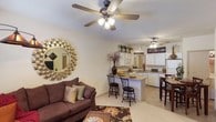 Located at 1800 Links Blvd in Tuscaloosa, Alabama, this single-story residence offers an open-concept layout that seamlessly connects the living area to the kitchen and dining space. The living room is a welcoming area anchored by a plush sofa, a decorative sunburst mirror, and a ceiling fan with integrated lighting that enhances comfort and airflow. Light-toned flooring runs throughout the space, contributing to a bright and airy atmosphere.

The kitchen is well-appointed with white cabinetry extending to the upper walls, providing generous storage. A peninsula counter with bar-height seating serves as a natural dividing element between the kitchen and living areas, making it ideal for casual dining or entertaining. The kitchen also features a refrigerator, microwave, and additional counter space. A second ceiling fan is positioned in the kitchen-dining area, reinforcing the unit's consistent airflow features.

The open layout creates a sense of spaciousness, while warm earth-toned décor and natural lighting give the interior a comfortable, inviting character. The overall design prioritizes functionality and flow throughout the connected living spaces.