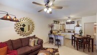 Located at 1800 Links Blvd in Tuscaloosa, Alabama, this single-story residence offers an open-concept layout that seamlessly connects the living area to the kitchen and dining space. The living room is a welcoming area anchored by a plush sofa, a decorative sunburst mirror, and a ceiling fan with integrated lighting that enhances comfort and airflow. Light-toned flooring runs throughout the space, contributing to a bright and airy atmosphere.

The kitchen is well-appointed with white cabinetry extending to the upper walls, providing generous storage. A peninsula counter with bar-height seating serves as a natural dividing element between the kitchen and living areas, making it ideal for casual dining or entertaining. The kitchen also features a refrigerator, microwave, and additional counter space. A second ceiling fan is positioned in the kitchen-dining area, reinforcing the unit's consistent airflow features.

The open layout creates a sense of spaciousness, while warm earth-toned décor and natural lighting give the interior a comfortable, inviting character. The overall design prioritizes functionality and flow throughout the connected living spaces.