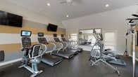 The fitness center at 31 Putnam Green in Greenwich, Connecticut is a well-appointed, single-floor exercise facility designed to support a comprehensive workout routine. The space features vaulted ceilings that lend an open, airy feel, complemented by recessed lighting and a ceiling fan that keeps the environment comfortable during workouts. A distinctive horizontal striped accent wall in gold and white adds a modern, energetic aesthetic throughout the room.

The cardio zone is well-equipped with a row of Star Trac treadmills, each featuring individual digital displays, alongside elliptical trainers and a stationary cycle — all positioned facing wall-mounted flat-screen televisions for an engaging workout experience. A large mirror along one wall visually expands the space and allows users to monitor their form.

A dedicated free weights area occupies a naturally lit corner of the facility, featuring a full dumbbell rack with a wide range of weights, exercise mats, a stability ball, and additional fitness accessories. Framed artwork decorates the walls in this section, adding a polished touch.

Large windows throughout the space flood the interior with natural light and offer views of an outdoor deck and surrounding greenery, creating an inviting and motivating atmosphere. Rubber flooring throughout ensures safety and durability across all workout zones.