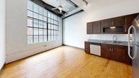 This 1-bedroom, 1-bathroom loft unit at 101 E Sixth St in downtown Winston-Salem showcases industrial character with soaring ceilings, exposed steel beams, and warm original hardwood floors throughout. The open living and kitchen area is flooded with natural light through large, multi-pane factory-style windows. The kitchen features dark wood cabinetry, granite countertops, and stainless steel appliances, including a range, refrigerator, and dishwasher. Track lighting and a ceiling fan complete the space. The bedroom offers hardwood floors and a transom window, while the bathroom includes a combination tub and shower. A hallway and closet round out this well-appointed urban loft.