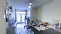 This single-floor space offers a warm and inviting atmosphere, thoughtfully arranged to maximize comfort and functionality. The living area serves as the heart of the home, highlighted by a ceiling fan that keeps the space airy and comfortable. A striking exposed brick accent wall adds a touch of historic character and visual texture, complementing the neutral-toned walls throughout.

Natural light pours generously into the room through a set of classic French doors at the front of the space, creating a bright and welcoming entry point. The doors are framed by the charming brick surround, reinforcing the unit's blend of traditional and cozy aesthetics.

The living area is well-furnished with a comfortable sofa adorned with accent pillows, a wooden coffee table, and a patterned area rug that anchors the seating arrangement. A dining area is positioned nearby, with a table and chairs that flow naturally from the main living space, making the layout well-suited for both relaxing and entertaining.

Additional storage is provided by a dresser tucked near the entryway, adding practical utility without interrupting the room's open feel. Overall, the space exudes a lived-in warmth, combining classic architectural details with a functional, homey layout.