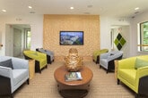 This contemporary single-floor lounge space is thoughtfully designed to blend comfort, style, and functionality. The entrance features floor-to-ceiling black-framed glass doors that flood the interior with natural light and provide views of lush greenery beyond.

The lounge is anchored by a striking textured accent wall composed of geometric, three-dimensional panels in a warm neutral tone, serving as the backdrop for a wall-mounted flat-screen TV. In front of it, a curated seating arrangement features modern curved armchairs in chartreuse and grey tones, complemented by a round wooden coffee table and natural wood stump accents, all set atop a woven area rug that adds warmth and texture.

Adjacent to the lounge area, a communal gathering zone centers around a substantial live-edge wood table with dark metal-framed bar stools, ideal for collaborative or social use. Overhead, an eye-catching cluster of globe pendant lights in varying heights creates an inviting and design-forward atmosphere. Built-in cabinetry with stainless steel appliances lines the wall nearby, adding utility to the space.

White tile flooring, recessed lighting, and crisp white walls throughout maintain a clean, open aesthetic. A decorative moss panel art installation adds a vibrant natural element. Large windows and glass doors ensure the space remains bright and connected to the outdoors.
