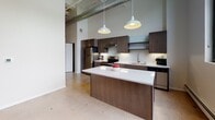 Located at 128 N Washington Ave in Minneapolis's vibrant North Loop neighborhood, this single-floor unit blends industrial character with contemporary finishes throughout. Polished concrete floors run continuously across the open layout, anchoring the space with an urban aesthetic that complements the building's architectural heritage.

The kitchen serves as a central focal point, featuring dark espresso cabinetry, stainless steel appliances, a subway tile backsplash, and a generous white quartz island beneath a pair of pendant lights. Soaring ceilings with exposed ductwork add loft-like volume to the cooking and living area, while large black-framed windows frame views of the Minneapolis skyline and street below.

The bedroom showcases exposed yellow brick along an upper accent wall, lending warmth and historic character to the space. Two sets of double closet doors provide ample storage, while additional oversized windows flood the room with natural light and urban views. A baseboard heating system runs along the perimeter.

The bathroom offers a refined finish package, including a walk-in shower clad in marble-patterned tile with sliding glass doors, dark wood vanity cabinetry, a white quartz countertop, and wood-look plank flooring. The overall ambiance throughout is clean, modern, and distinctly urban.
