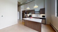 Located at 128 N Washington Ave in Minneapolis's vibrant North Loop neighborhood, this single-floor unit blends industrial character with contemporary finishes throughout. Polished concrete floors run continuously across the open layout, anchoring the space with an urban aesthetic that complements the building's architectural heritage.

The kitchen serves as a central focal point, featuring dark espresso cabinetry, stainless steel appliances, a subway tile backsplash, and a generous white quartz island beneath a pair of pendant lights. Soaring ceilings with exposed ductwork add loft-like volume to the cooking and living area, while large black-framed windows frame views of the Minneapolis skyline and street below.

The bedroom showcases exposed yellow brick along an upper accent wall, lending warmth and historic character to the space. Two sets of double closet doors provide ample storage, while additional oversized windows flood the room with natural light and urban views. A baseboard heating system runs along the perimeter.

The bathroom offers a refined finish package, including a walk-in shower clad in marble-patterned tile with sliding glass doors, dark wood vanity cabinetry, a white quartz countertop, and wood-look plank flooring. The overall ambiance throughout is clean, modern, and distinctly urban.