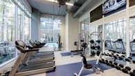 Located at 2610 Erwin Road in Durham, NC, this modern fitness center is thoughtfully designed to support a full range of workout routines in a bright, energizing environment. The space features soaring ceilings with exposed ductwork, lending an industrial-chic aesthetic that pairs well with the clean, contemporary finishes throughout. Large floor-to-ceiling windows flood the interior with abundant natural light and offer direct views of an outdoor pool area, creating a motivating and visually connected workout experience. A large ceiling fan overhead ensures comfortable air circulation throughout the space. The fitness floor is well-equipped with a lineup of treadmills and other cardio machines along the windowed wall, complemented by a comprehensive selection of strength training equipment, including cable machines, weight benches, and a free weight rack. Blue accent mats define designated workout zones, adding a pop of color to the light-toned flooring. Full-length mirrors along one wall enhance the sense of spaciousness while allowing users to monitor their form. The overall ambiance is clean, open, and purposefully laid out, making this fitness center a well-appointed amenity for the surrounding community.