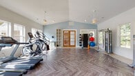Located in Acworth, Georgia, this single-floor fitness center offers a thoughtfully designed workout space that combines functionality with an inviting aesthetic. The facility is characterized by soaring vaulted ceilings that create an open, airy atmosphere, complemented by soft blue accent walls and crisp white trim throughout.

The space is divided into distinct workout zones. The cardio area features multiple treadmills, elliptical machines, a stationary bike, and a rowing machine, all arranged along a wall of large, multi-pane windows that flood the room with natural light and offer views of the surrounding greenery. Elegant pendant ceiling fans provide both ventilation and visual appeal.

The strength training zone is equally well-appointed, housing a selection of weight machines, a dumbbell rack, adjustable benches, and a cable/functional trainer system. Storage racks along the wall hold a variety of exercise balls and resistance accessories, while open shelving units provide convenient organization for additional equipment and personal items.

Underfoot, a striking herringbone-patterned luxury vinyl floor ties the entire space together, offering both durability and visual sophistication. Natural wood-framed double French doors serve as a central architectural focal point, leading out from the main exercise area. A compact beverage refrigerator adds a convenient amenity for users. The facility is bright, clean, and comprehensively equipped for a full range of fitness activities.