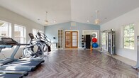 Located in Acworth, Georgia, this single-floor fitness center offers a thoughtfully designed workout space that combines functionality with an inviting aesthetic. The facility is characterized by soaring vaulted ceilings that create an open, airy atmosphere, complemented by soft blue accent walls and crisp white trim throughout.

The space is divided into distinct workout zones. The cardio area features multiple treadmills, elliptical machines, a stationary bike, and a rowing machine, all arranged along a wall of large, multi-pane windows that flood the room with natural light and offer views of the surrounding greenery. Elegant pendant ceiling fans provide both ventilation and visual appeal.

The strength training zone is equally well-appointed, housing a selection of weight machines, a dumbbell rack, adjustable benches, and a cable/functional trainer system. Storage racks along the wall hold a variety of exercise balls and resistance accessories, while open shelving units provide convenient organization for additional equipment and personal items.

Underfoot, a striking herringbone-patterned luxury vinyl floor ties the entire space together, offering both durability and visual sophistication. Natural wood-framed double French doors serve as a central architectural focal point, leading out from the main exercise area. A compact beverage refrigerator adds a convenient amenity for users. The facility is bright, clean, and comprehensively equipped for a full range of fitness activities.