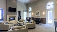 This single-floor space presents an elegantly designed lounge or common area that blends classic architectural details with modern comfort. The room immediately draws attention to its soaring ceilings, which are accentuated by crisp crown molding and warm neutral-toned walls featuring dark wood wainscoting along the lower half — a refined design detail that adds depth and sophistication throughout.

Natural light pours in through tall, arched windows, providing views of lush greenery outside. A mix of seating arrangements caters to various needs: a casual lounge area offers plush, neutral-upholstered armchairs and sofas gathered around a round dark wood coffee table, while a high-top bar-style seating area near the windows provides a more upright, social setting with dark wood chairs and tables.

A fireplace anchors one wall, contributing to the warm, inviting ambiance of the space, while a wall-mounted flat-screen television nearby adds a functional entertainment element. Built-in shelving adjacent to the fireplace offers additional visual interest. Framed abstract artwork and wall-mounted sconce lighting further enhance the polished, welcoming atmosphere of this thoughtfully appointed space.