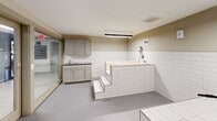 Located within the Rime Village community at 6200 Rime Village Dr NW in Huntsville, Alabama, this purpose-built pet grooming facility offers a clean, well-equipped space designed specifically for pet care. The room is finished with durable white subway tile wainscoting along the lower walls and small-format floor tiling throughout, providing a hygienic and easy-to-clean environment. At the center of the space stands a raised, step-access bathing tub constructed with tiled surround walls, accompanied by a mounted showerhead and handheld spray fixture for thorough pet washing. Gray shaker-style cabinetry with a dark countertop lines one wall, offering ample storage for grooming supplies, while a utility sink provides added convenience. Overhead, linear LED strip lighting ensures bright, even illumination across the entire space. A built-in ventilation unit is mounted to the ceiling, maintaining air quality and comfort. The room is accessed through full-height glass-panel sliding doors, which allow natural visibility from the corridor and lend a modern, transparent aesthetic to the entry. The overall design is functional and straightforward, prioritizing cleanliness, accessibility, and ease of use for residents and their pets.