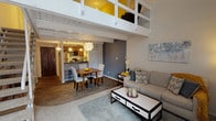 Located along Lindell Boulevard in the heart of St. Louis, this two-story loft-style model unit showcases a thoughtfully designed open-concept layout that blends modern living with architectural character. The main level features rich hardwood-style flooring, exposed white-painted beam ceilings, and a seamless flow between the living, dining, and kitchen areas. The living room is anchored by soaring double-height ceilings and a large window that invites abundant natural light, offering views of the surrounding streetscape. A compact, well-appointed kitchen includes warm wood cabinetry, dark granite-style countertops, and stainless steel appliances, including a range and over-the-range microwave. The dining area connects naturally to both the kitchen and living spaces, making it well-suited for entertaining. A staircase leads to the upper mezzanine level, where the bedroom retreat features plush carpeting, a bold yellow accent wall, and an open-railing overlook to the floor below. A second bedroom on the lower level also offers carpeted flooring and ample space. The bathroom is clean and functional, featuring a single-sink vanity with warm wood cabinetry. Throughout the unit, a cohesive color palette of grey, blue, and yellow accents creates a modern and inviting atmosphere.