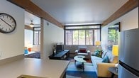 This single-floor space showcases a thoughtfully designed open-plan layout that seamlessly integrates a living area and sleeping zone. A structural wood-accented beam serves as a subtle architectural divider between the two areas, adding warmth and character to the otherwise modern aesthetic. The living area is anchored by a light gray sofa adorned with colorful accent pillows, complemented by a matching armchair and a contemporary curved coffee table. A vibrant, multicolored area rug grounds the seating arrangement and adds a lively pop of color to the space. A media console with a flat-screen TV sits against one wall, providing a functional entertainment setup. Large, floor-to-ceiling windows span the far wall, flooding the interior with abundant natural light and framing views of surrounding greenery and exterior architecture. The sleeping area, partially visible through the open partition, features a neatly made bed and coordinating lamp. Smooth white ceilings and light-toned walls maintain a clean, airy atmosphere throughout, while carefully placed lamps ensure warm, layered lighting. The overall ambiance is modern, vibrant, and welcoming.