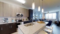 This stylish single-floor residence offers a thoughtfully designed open-concept layout that seamlessly connects the kitchen and living areas, creating an inviting and functional living environment. The kitchen is a standout feature, showcasing expansive granite countertops with subtle speckled detailing, a large undermount stainless steel sink, and a full suite of stainless steel appliances, including a range, oven, and over-the-range microwave. A two-tone cabinetry scheme pairs light wood upper cabinets with sleek dark lower cabinets, lending the kitchen a contemporary and sophisticated aesthetic. A peninsula island with pendant lighting overhead provides additional prep space and casual seating, making it ideal for entertaining. The open flow into the living area is highlighted by soft blue accent walls and large windows that allow natural light to fill the space, creating a bright and airy ambiance. The living area offers ample room for comfortable furnishings and stylish décor. Throughout the space, modern design choices — from the clean lines of the cabinetry to the polished fixtures — come together to deliver a cohesive and upscale residential experience on a single, well-appointed floor.