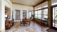 This single-floor community or leasing office is designed with a warm, inviting aesthetic that balances functionality with comfort. The space is unified by earthy, neutral wall tones, rich wood trim accents, and a multi-toned terracotta and beige tile floor that flows throughout all areas.

Natural light is a defining feature of this space. Large windows fitted with wood-slat blinds line the walls, while transom windows positioned above the main entrance doors draw in additional overhead light, creating a bright and open atmosphere. The tall ceilings, accented by bold crown molding with gold-toned trim, add a sense of height and grandeur.

The main entry area opens into a casual seating and meeting zone, furnished with a round wooden dining table and matching chairs — an ideal setting for informal conversations or small group gatherings. Adjacent to this is a dedicated workspace featuring a rectangular desk with woven accent chairs, framed by views of the surrounding landscape through wide windows.

A formal office area is also present, equipped with a full-size desk and chairs to accommodate one-on-one meetings or administrative work. Decorative touches, including potted plants, floral arrangements, framed artwork, and a console table with curated accessories, contribute to the polished yet welcoming ambiance throughout the space.